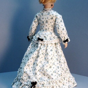 K&ouml;nnte beinhalten: Ein wei&szlig;es Kleid mit Blumenmuster und schwarzen Schleifenakzenten. Das Kleid hat ein tailliertes Oberteil und einen vollen Rock mit R&uuml;schen.