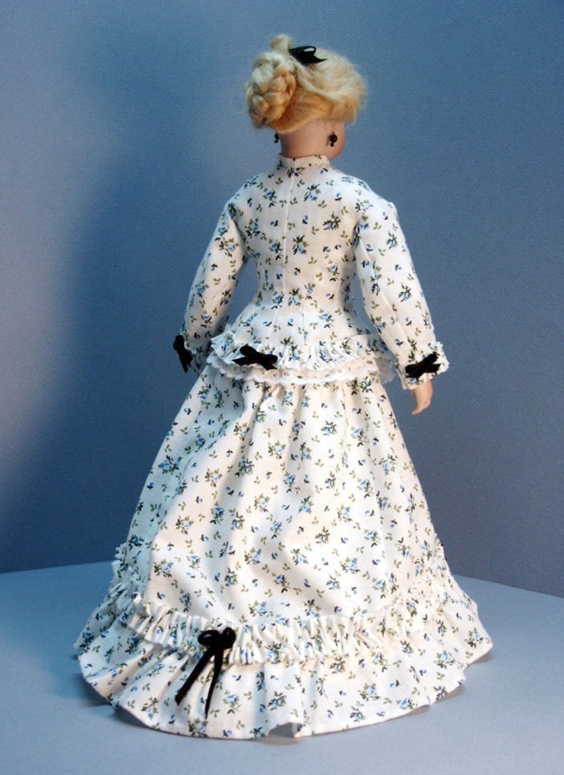K&ouml;nnte beinhalten: Ein wei&szlig;es Kleid mit Blumenmuster und schwarzen Schleifenakzenten. Das Kleid hat ein tailliertes Oberteil und einen vollen Rock mit R&uuml;schen.