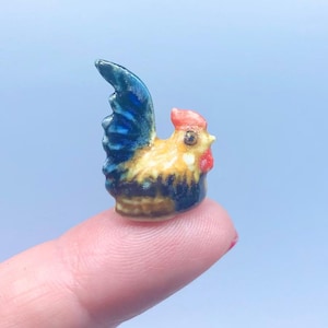 May include: A small ceramic rooster figurine with a blue and black tail, yellow and brown body, and a red comb.
