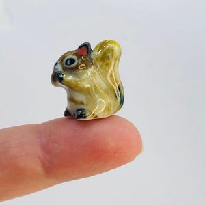 May include: A small ceramic squirrel figurine with brown, white, and yellow paint. The squirrel is sitting on its hind legs with its tail curled up behind it.