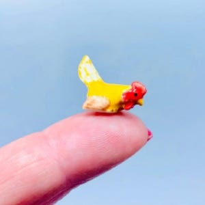 May include: A small ceramic chicken figurine, predominantly yellow with a red comb and wattle. The miniature sculpture is resting on a fingertip against a light blue backdrop. The chicken has a detailed design and is likely a decorative item.