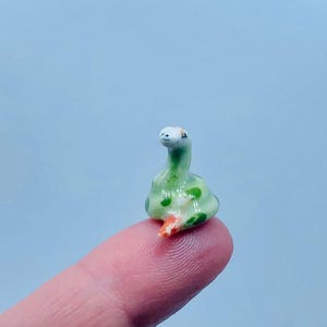 May include: A small ceramic figurine of a green snake with orange and white accents. The snake is curled up in a sitting position with its head tilted to the side.