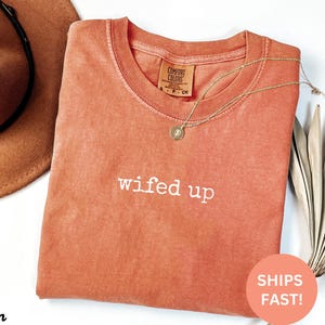 Puede incluir: Una camiseta Comfort Colors de color coral con la frase bordada "wifed up". Un collar de oro con un colgante descansa sobre la camisa. Un sombrero marr&oacute;n es visible en la esquina superior izquierda. Una pegatina que dice "SHIPS FAST!" est&aacute; en la esquina inferior derecha.