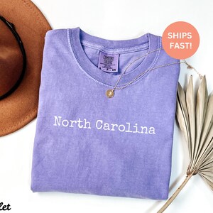 Op de afbeelding: Een paarskleurig T-shirt met de woorden "North Carolina" in het wit. Een gouden ketting met een hanger is over het shirt gedrapeerd. Een bruine hoed en gedroogde palmbladeren zijn op de achtergrond.