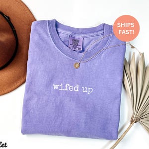Puede incluir: Una camiseta de color violeta con la frase "wifed up" bordada en blanco. Un collar dorado con un colgante est&aacute; sobre la camiseta. Un sombrero marr&oacute;n y una hoja de palma seca est&aacute;n en el fondo.