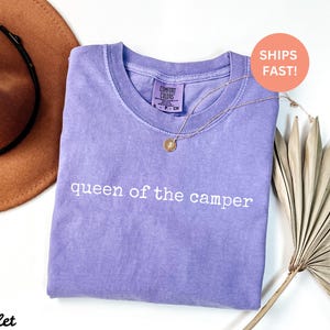 May include: A violet-colored t-shirt with the text "queen of the camper" in white. A gold necklace with a pendant is on the shirt. A brown hat and dried palm leaf are in the background.