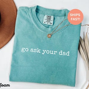 May include: Seafoam green t-shirt with the phrase "go ask your dad" in white lettering. The shirt has a crew neck and a Comfort Colors tag. A gold necklace with a pendant is draped across the shirt.