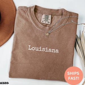 May include: Espresso-colored t-shirt with the word "Louisiana" in white block letters. A silver necklace with a small pendant rests on the shirt. The shirt is folded, and a brown hat is visible in the upper left corner.