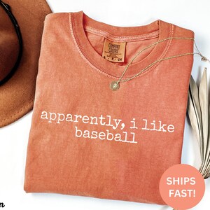 May include: A coral-coloured t-shirt with the phrase "apparently, i like baseball" in white lettering. A gold necklace with a pendant rests on the shirt. A brown hat is visible in the upper left corner.