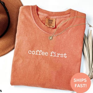 Puede incluir: Una camiseta Comfort Colors de color coral con la frase "coffee first" en letras blancas. Un collar dorado con un colgante descansa sobre la camiseta. Un sombrero marr&oacute;n es visible en la esquina superior izquierda.