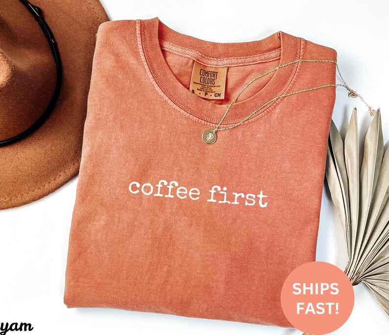 Puede incluir: Una camiseta Comfort Colors de color coral con la frase "coffee first" en letras blancas. Un collar dorado con un colgante descansa sobre la camiseta. Un sombrero marr&oacute;n es visible en la esquina superior izquierda.