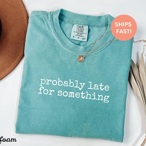 Puede incluir: Camiseta verde mar con el texto blanco "probably late for something". La camiseta tiene cuello redondo y mangas cortas. Un collar dorado con un colgante est&aacute; sobre la camiseta.