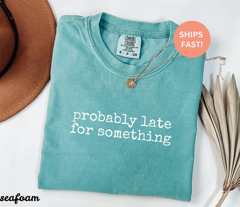 Puede incluir: Camiseta verde mar con el texto blanco "probably late for something". La camiseta tiene cuello redondo y mangas cortas. Un collar dorado con un colgante est&aacute; sobre la camiseta.
