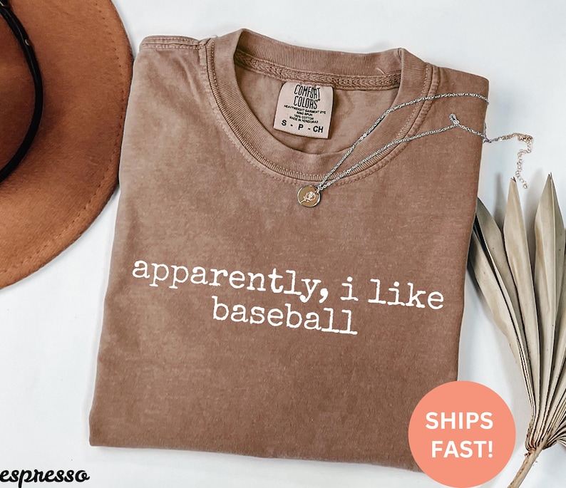 May include: Brown t-shirt with the white text "apparently, i like baseball". The shirt has a crew neck and a small label on the upper chest. A silver necklace with a pendant is draped across the shirt.