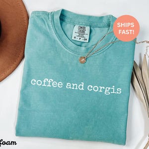 Puede incluir: Camiseta verde mar con las palabras "coffee and corgis" en blanco. Un collar dorado con un colgante descansa sobre la camiseta. La camiseta est&aacute; doblada y junto a un sombrero marr&oacute;n y una planta seca.