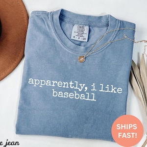 May include: A blue jean coloured t-shirt with the text "apparently, i like baseball" in white. A gold necklace with a pendant is draped across the shirt. The shirt is folded on a white surface.