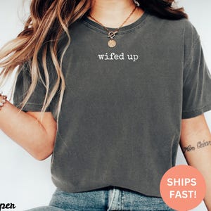 Puede incluir: Camiseta gris oscuro con el texto blanco "wifed up". La camiseta es de cuello redondo y parece estar hecha de un material suave y c&oacute;modo. La camiseta se usa con vaqueros azules.
