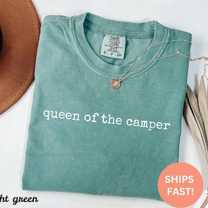 May include: A light green t-shirt with the text "queen of the camper" in white. The shirt has a crew neck and short sleeves. A gold necklace with a pendant is on the shirt.