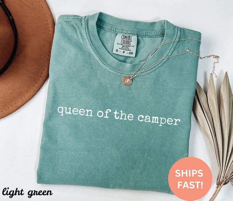 May include: A light green t-shirt with the text "queen of the camper" in white. The shirt has a crew neck and short sleeves. A gold necklace with a pendant is on the shirt.