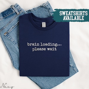 May include: A navy blue t-shirt with the white text "brain loading... please wait". The t-shirt is folded on top of blue denim jeans. A teal box in the upper right corner reads "SWEATSHIRTS AVAILABLE".