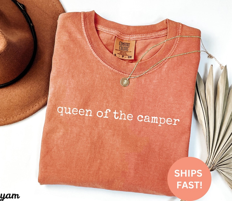 May include: A coral-colored Comfort Colors t-shirt with the phrase "queen of the camper" in white lettering. A gold necklace with a pendant rests on the shirt. A brown hat is visible in the upper left corner. A sticker that says "SHIPS FAST!" is in the lower right corner.