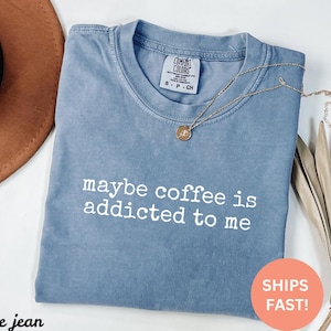 May include: A blue jean Comfort Colors t-shirt with the phrase "maybe coffee is addicted to me" in white. A gold necklace with a pendant is draped across the shirt. The shirt is folded on a white surface.