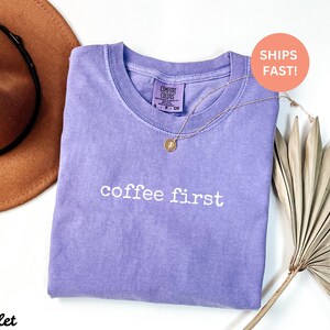Puede incluir: Una camiseta violeta con la frase "coffee first" en letras blancas. Un collar dorado con un colgante descansa sobre la camiseta. Un sombrero marr&oacute;n y una hoja de palma seca est&aacute;n en el fondo.