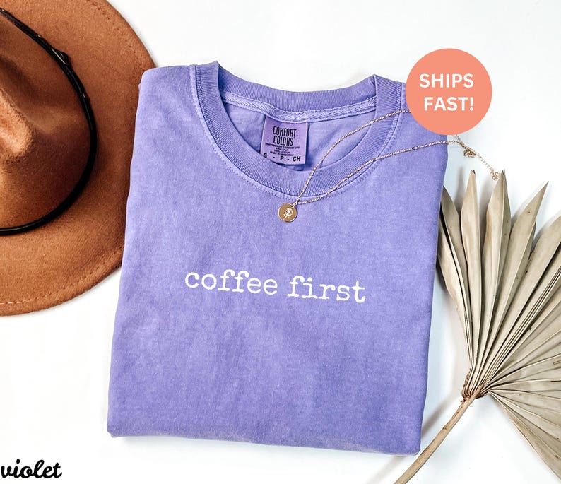 Puede incluir: Una camiseta violeta con la frase "coffee first" en letras blancas. Un collar dorado con un colgante descansa sobre la camiseta. Un sombrero marr&oacute;n y una hoja de palma seca est&aacute;n en el fondo.