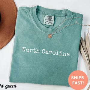Op de afbeelding: Lichtgroen T-shirt met de tekst "North Carolina" in het wit. Het shirt heeft een ronde hals en korte mouwen. Een gouden ketting met een hanger is over het shirt gedrapeerd. Het shirt is op een wit oppervlak gevouwen.