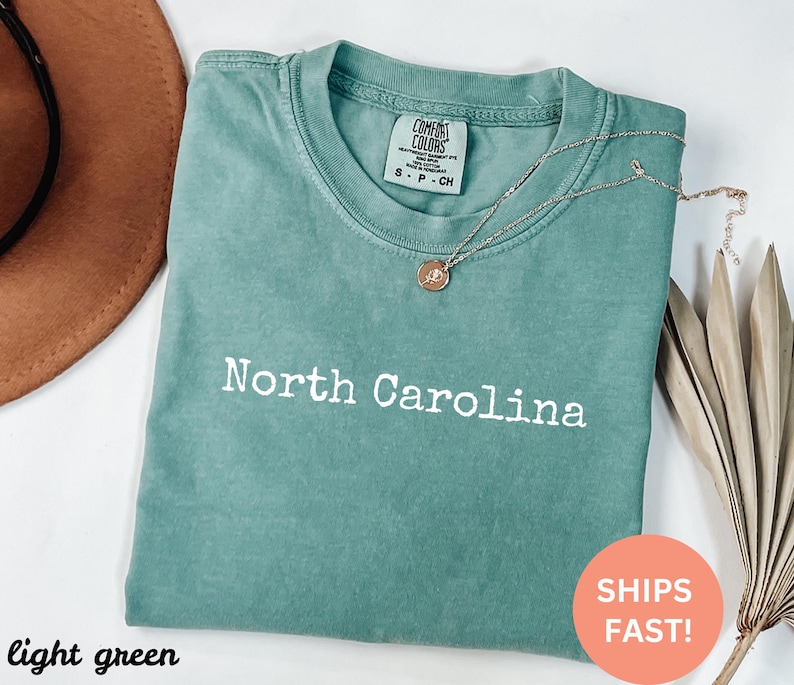Op de afbeelding: Lichtgroen T-shirt met de tekst "North Carolina" in het wit. Het shirt heeft een ronde hals en korte mouwen. Een gouden ketting met een hanger is over het shirt gedrapeerd. Het shirt is op een wit oppervlak gevouwen.