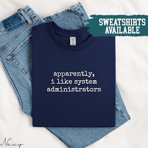 Könnte beinhalten: Marineblaues T-Shirt mit dem weißen Text "apparently, i like system administrators". Das T-Shirt ist auf einer Jeans gefaltet. Ein türkisfarbenes Banner oben rechts lautet "SWEATSHIRTS AVAILABLE".