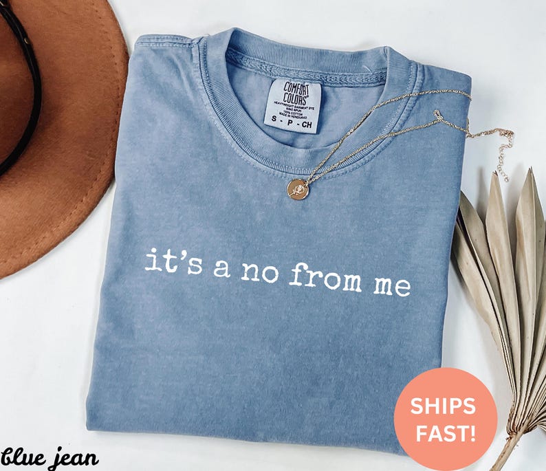 May include: A blue jean Comfort Colors t-shirt with the phrase "it's a no from me" printed in white. A gold necklace with a pendant is draped across the shirt. The shirt is folded and ready to wear.