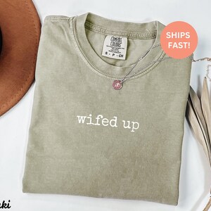 Puede incluir: Camiseta de color caqui con el texto blanco "wifed up". La camiseta tiene cuello redondo y mangas cortas. Un collar plateado con un colgante rosa est&aacute; drapeado sobre la camiseta. La camiseta est&aacute; doblada y colocada sobre una superficie blanca.