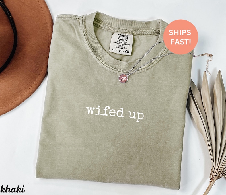 Puede incluir: Camiseta de color caqui con el texto blanco "wifed up". La camiseta tiene cuello redondo y mangas cortas. Un collar plateado con un colgante rosa est&aacute; drapeado sobre la camiseta. La camiseta est&aacute; doblada y colocada sobre una superficie blanca.