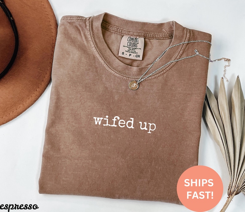 Puede incluir: Camiseta marr&oacute;n con la frase "wifed up" en letras blancas. Un collar plateado con un peque&ntilde;o colgante descansa sobre la camiseta. La camiseta est&aacute; doblada y un sombrero marr&oacute;n es visible en el fondo.