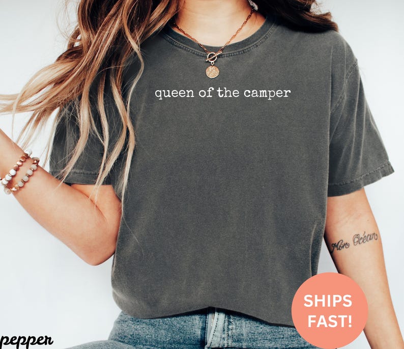 May include: Dark gray t-shirt with the text "queen of the camper" in white. The shirt is a crew neck style and has short sleeves. The shirt is made of a soft material.