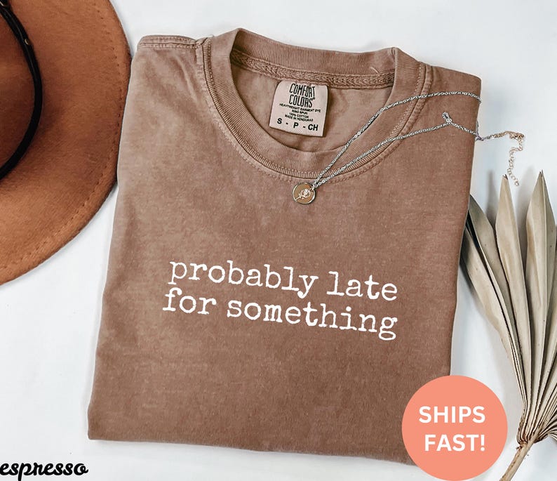 Puede incluir: Camiseta marr&oacute;n con la frase "probably late for something" en letras blancas. Un collar plateado con un peque&ntilde;o colgante descansa sobre la camiseta. Un sombrero marr&oacute;n y una planta seca son visibles en el fondo. La camiseta est&aacute; doblada.
