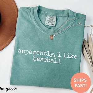 May include: Light green t-shirt with the text "apparently, i like baseball" in white. The shirt has a crew neck and short sleeves. A gold necklace with a pendant is draped across the shirt.