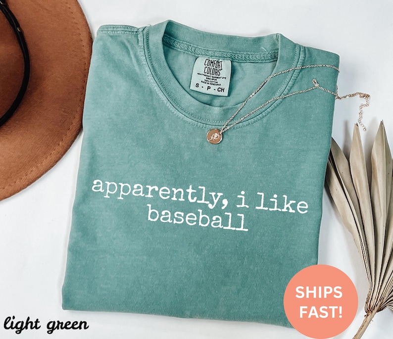 May include: Light green t-shirt with the text "apparently, i like baseball" in white. The shirt has a crew neck and short sleeves. A gold necklace with a pendant is draped across the shirt.