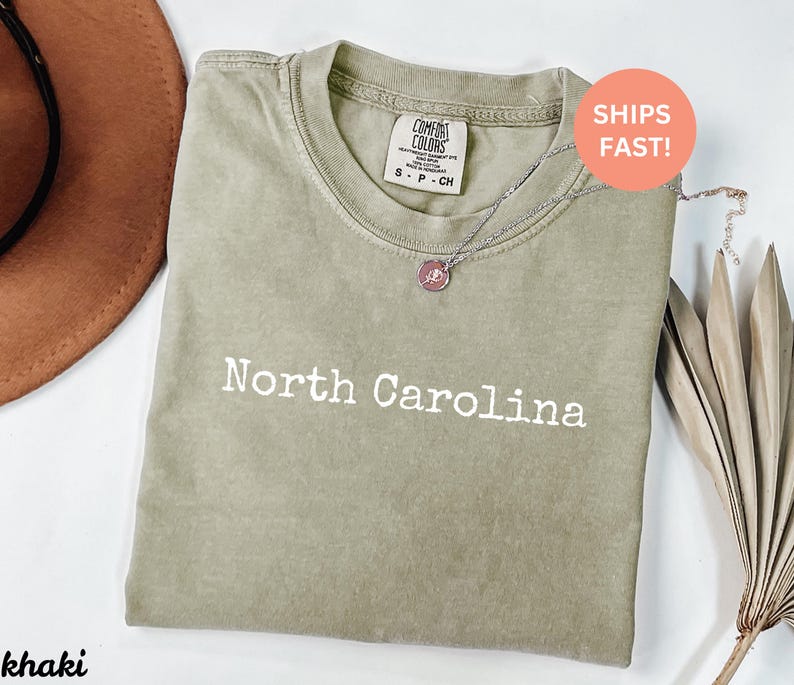 Op de afbeelding: Khaki-kleurig T-shirt met de woorden "North Carolina" in het wit gedrukt. Het shirt heeft een ronde hals en korte mouwen. Een zilveren ketting met een hanger is over het shirt gedrapeerd. Het shirt is op een wit oppervlak gevouwen.