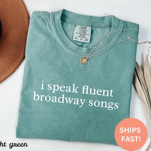 May include: Light green t-shirt with the text "i speak fluent broadway songs" in white. A gold necklace with a pendant rests on the shirt. The shirt is folded and has a Comfort Colors tag.