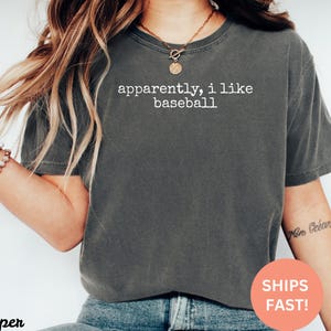 May include: Dark grey t-shirt with the text "apparently, i like baseball" in white. The shirt is a crew neck and has short sleeves. The image also includes a bracelet and a necklace.