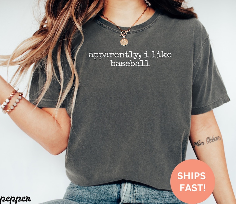 May include: Dark grey t-shirt with the text "apparently, i like baseball" in white. The shirt is a crew neck and has short sleeves. The image also includes a bracelet and a necklace.