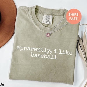 May include: Khaki-coloured t-shirt with the text "apparently, i like baseball" in white. The shirt has a crew neck and a Comfort Colors tag. A silver necklace with a pendant is draped across the shirt.