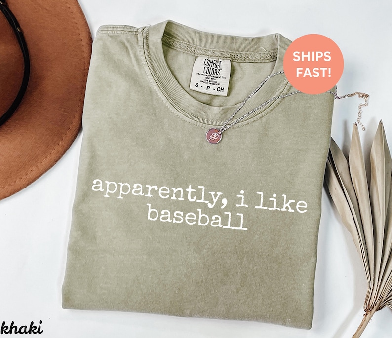 May include: Khaki-coloured t-shirt with the text "apparently, i like baseball" in white. The shirt has a crew neck and a Comfort Colors tag. A silver necklace with a pendant is draped across the shirt.