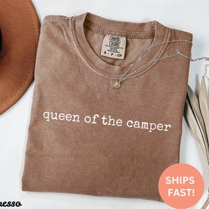 May include: Espresso-colored t-shirt with the text "queen of the camper" in white. A silver necklace with a small pendant rests on the shirt. A brown hat and dried plant are in the background.