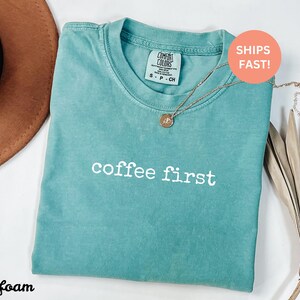 Puede incluir: Camiseta verde mar con la frase "coffee first" en blanco. La camiseta tiene cuello redondo y una peque&ntilde;a etiqueta en el cuello. Un collar dorado con un colgante est&aacute; sobre la camiseta. Un sombrero marr&oacute;n y hojas secas en el fondo.