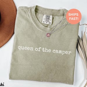 May include: Khaki-colored t-shirt with the text "queen of the camper" in white. The shirt has a crew neck and short sleeves. A silver necklace with a pendant is draped on the shirt. A brown hat is visible in the upper left corner.