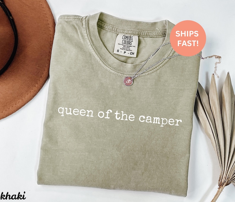 May include: Khaki-colored t-shirt with the text "queen of the camper" in white. The shirt has a crew neck and short sleeves. A silver necklace with a pendant is draped on the shirt. A brown hat is visible in the upper left corner.