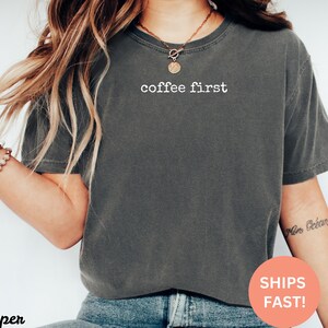 Puede incluir: Camiseta gris oscuro con el texto blanco "coffee first". La camiseta es de cuello redondo y est&aacute; hecha de un material suave. La camiseta se usa con vaqueros azules.